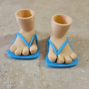 Könnte beinhalten: Zwei neuartige Pflanzgefäße in Form von Füßen mit blauen Flip-Flops. Die Füße sind hellbraun und die Flip-Flops haben ein schlichtes Design. Die Pflanzgefäße haben eine zylindrische Öffnung an der Oberseite, ideal für kleine Pflanzen oder Sukkulenten.