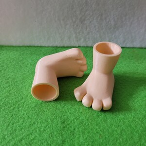 K&ouml;nnte beinhalten: Zwei 3D-gedruckte Puppenf&uuml;&szlig;e, einer mit einem ausgeh&ouml;hlten Kn&ouml;chel, der andere mit einem ausgeh&ouml;hlten Fu&szlig;. Die F&uuml;&szlig;e sind hellpfirsichfarben und befinden sich auf einer gr&uuml;nen Oberfl&auml;che.