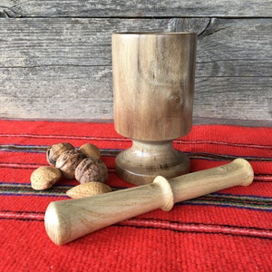 Puede incluir: Un mortero y un pilón de madera sobre una tela de rayas rojas y azules. El mortero es marrón y tiene una base redonda. El pilón también es marrón y tiene un extremo redondeado. Hay almendras y nueces esparcidas alrededor del mortero y el pilón.