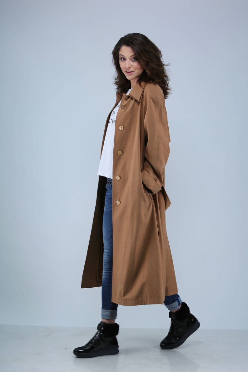 May include: A long, tan trench coat with large wooden buttons. The coat is worn over a white t-shirt and blue jeans. Black boots complete the outfit. The coat has a classic, timeless design.