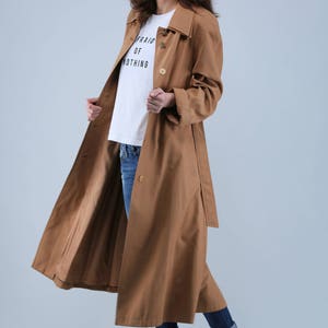 May include: A long, tan trench coat with a matching belt and button closure. The coat is worn over a white t-shirt with the text "AFRAID OF NOTHING" and blue jeans. Black boots complete the outfit.