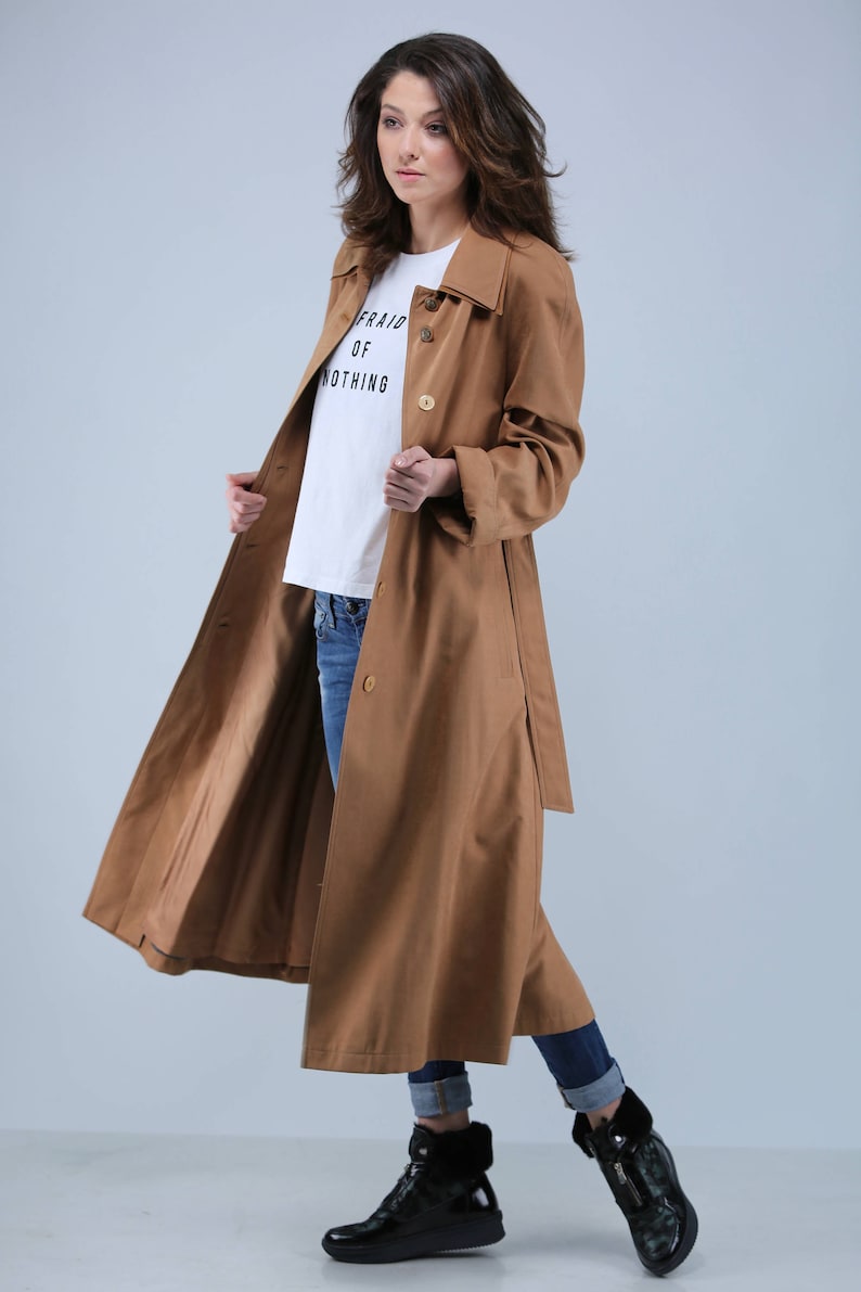 May include: A long, tan trench coat with a matching belt and button closure. The coat is worn over a white t-shirt with the text "AFRAID OF NOTHING" and blue jeans. Black boots complete the outfit.
