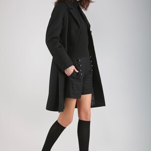 May include: A woman wearing a black coat, black shorts with button detailing, black knee-high socks, and black heels.