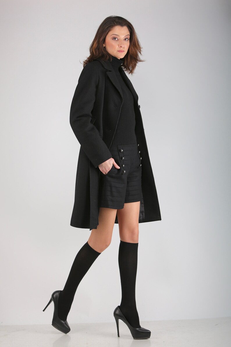 May include: A woman wearing a black coat, black shorts with button detailing, black knee-high socks, and black heels.
