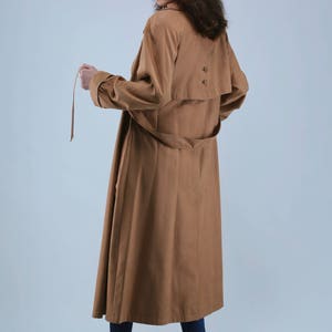 May include: A long, tan trench coat with a matching belt and button details. The coat has long sleeves and a flowing design. The model is wearing blue jeans and black boots. The coat is a classic outerwear piece.