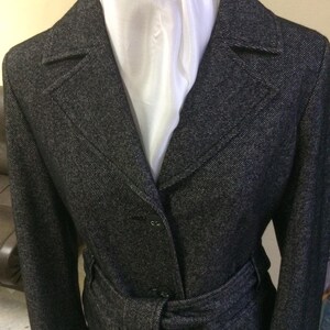 May include: A black and white tweed coat with a white scarf and a belt. The coat has a notched collar and a single-breasted closure.