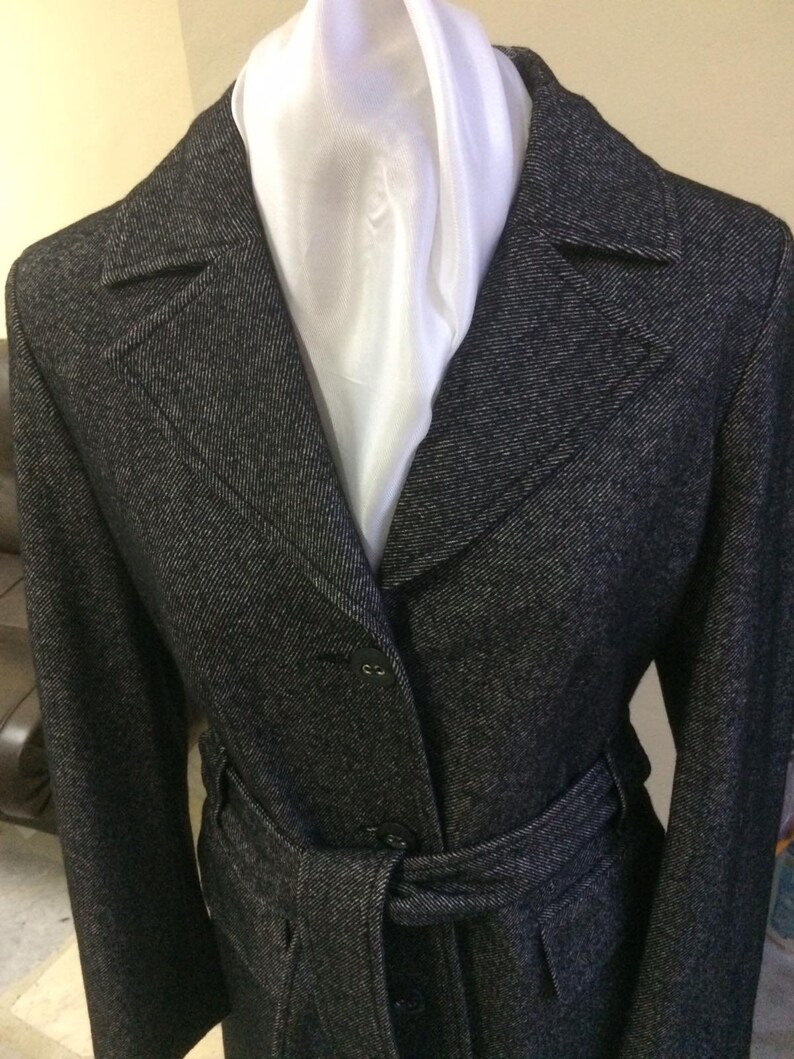 May include: A black and white tweed coat with a white scarf and a belt. The coat has a notched collar and a single-breasted closure.