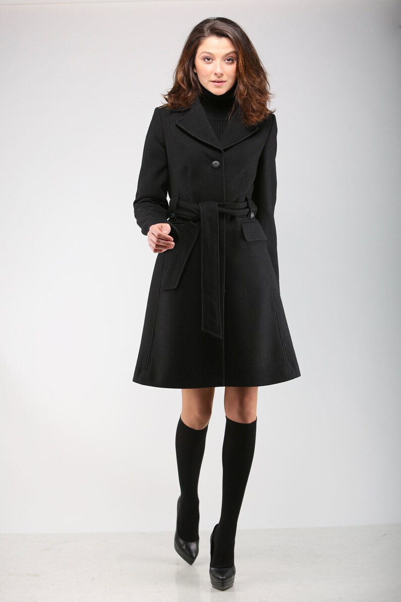 May include: A black belted coat with a mock neck collar. The coat is knee-length and has a fitted silhouette.