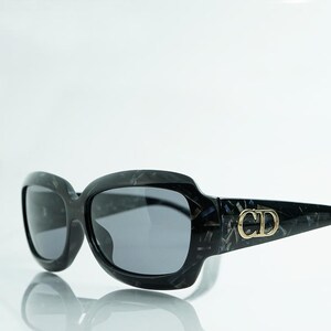 May include: Black and gray sunglasses with a marbled pattern. The sunglasses have a thick frame and dark lenses. The temples feature a gold-colored "CD" logo. The sunglasses are displayed on a reflective surface.