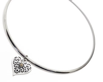 May include: A silver-coloured necklace with a heart-shaped pendant. The pendant has an openwork design and a small, circular accent. The necklace features a simple, rounded band.