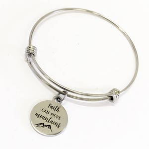 Puede incluir: Una pulsera de brazalete plateada con un dije redondo. El dije está grabado con las palabras "Faith can move mountains" y un gráfico de montaña. La pulsera es de metal brillante.