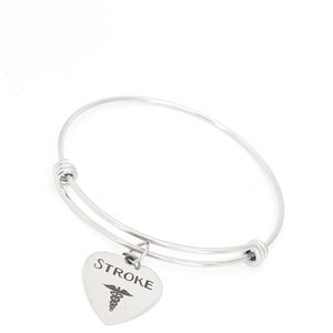 Puede incluir: Pulsera de brazalete de plata con un dije en forma de corazón que dice "STROKE" y tiene un símbolo médico grabado.