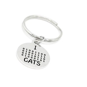 Può includere: Portachiavi d'argento con un ciondolo rotondo che dice "I  CATS" con una rappresentazione in codice binario della parola "CATS" attorno all'esterno del ciondolo.