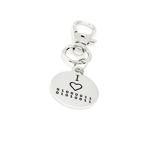 以下が含まれることがあります： 丸いタグに「I ♥ 01000011 01010100 01010011」と書かれたシルバーのキーホルダー。