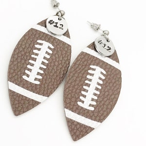 May include: A pair of brown leather football-shaped earrings with white stitching and a silver charm with the number "#12" on each earring.