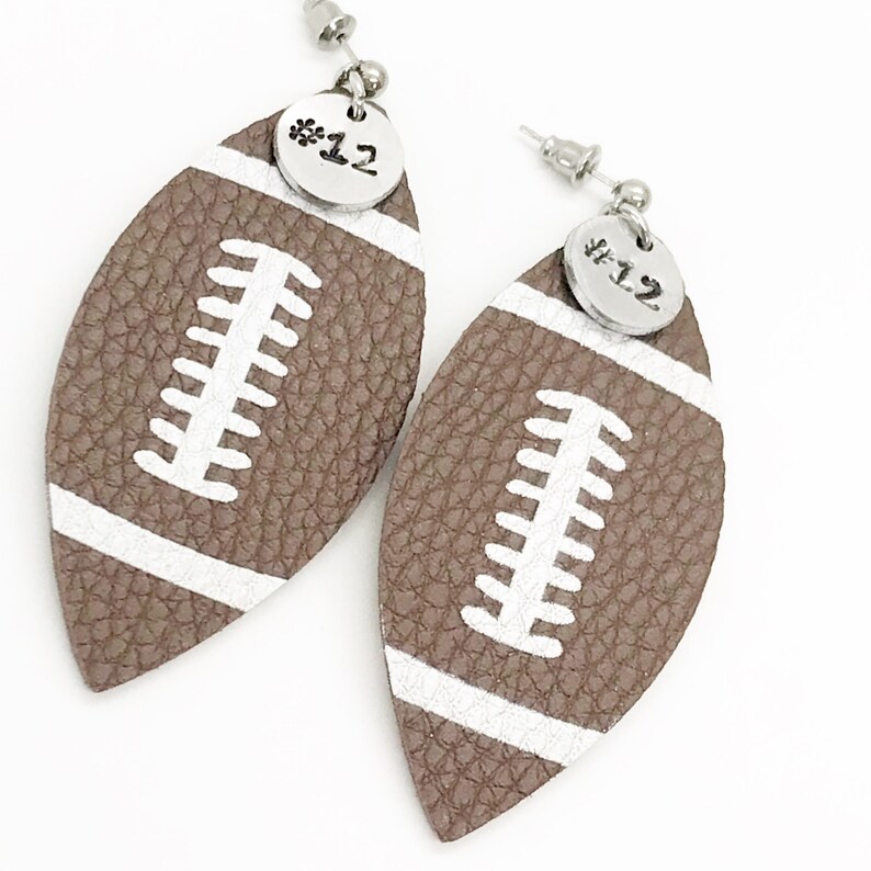 May include: A pair of brown leather football-shaped earrings with white stitching and a silver charm with the number "#12" on each earring.