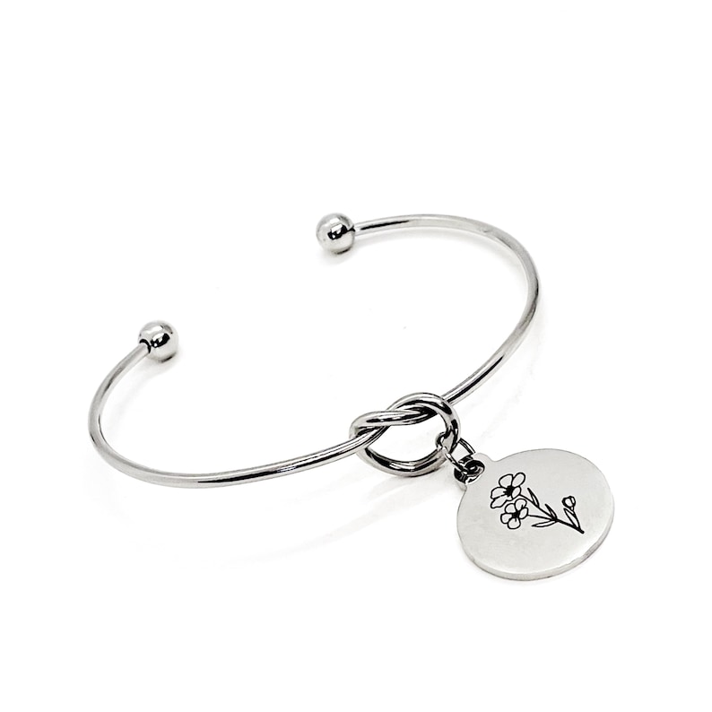 May include: Silver bangle bracelet with a knot detail and a round charm with a floral design.
