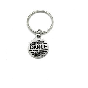 May include: A silver-coloured keyring with a round charm. The charm features the word "DANCE" in bold letters, with other words like "TEAM", "PERSEVERANCE", and "STRENGTH" around it. The keyring is set against a white background.