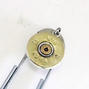 K&ouml;nnte beinhalten: Eine goldfarbene Schrotpatronenh&uuml;lse mit dem Text "WIN 20 GA" und einem silbernen Sprungring, der oben befestigt ist. Die H&uuml;lse ist an einer silbernen B&uuml;roklammer befestigt.