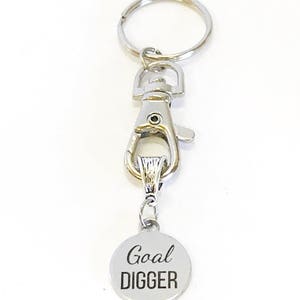 May include: Silver keychain with a silver clip and a round silver charm that says "Goal Digger" in black lettering.