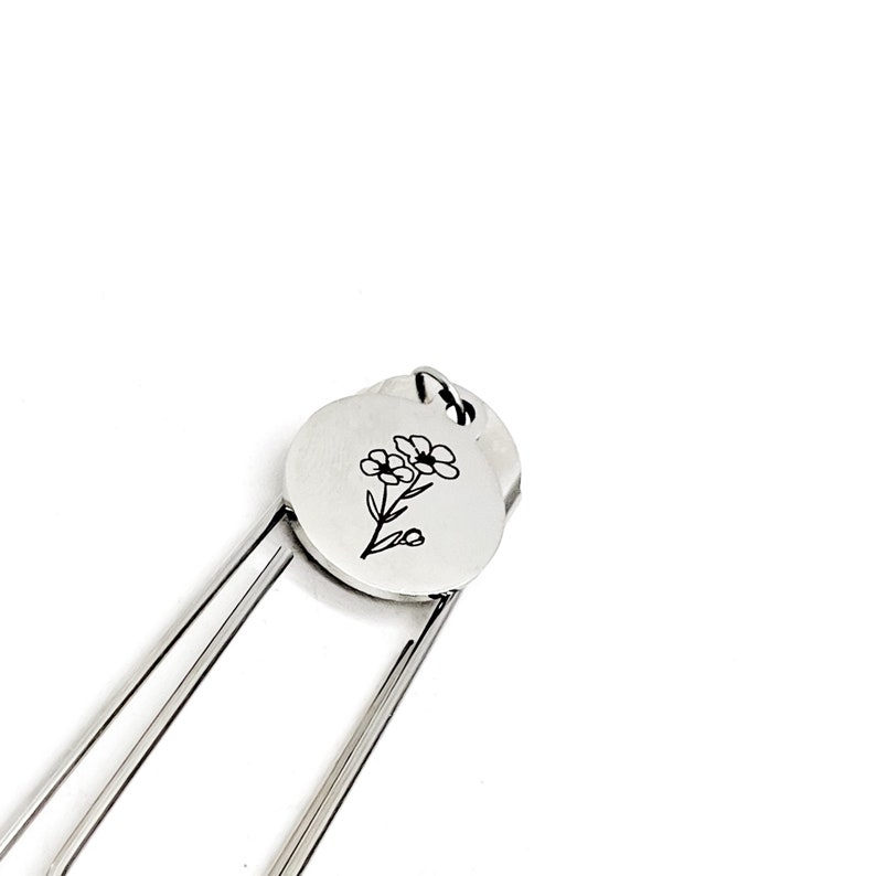 May include: Silver bookmark with a round charm featuring a simple line drawing of a flower.