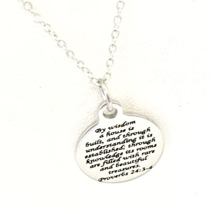 Pode incluir: Colar de prata com um pingente redondo que apresenta uma citação inspiradora de Provérbios 24:3-4: "Pela sabedoria se constrói uma casa, e pelo entendimento se firma; pelo conhecimento seus aposentos se enchem de tesouros preciosos e belos."