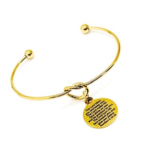 May include: Gold knot bangle bracelet with a round charm that has a bible verse inscribed on it. The verse reads: "The Lord bless you and keep you. The Lord make His face shine on you and be gracious to you. The Lord turn His face toward you and give you peace. Numbers 6:24-26"