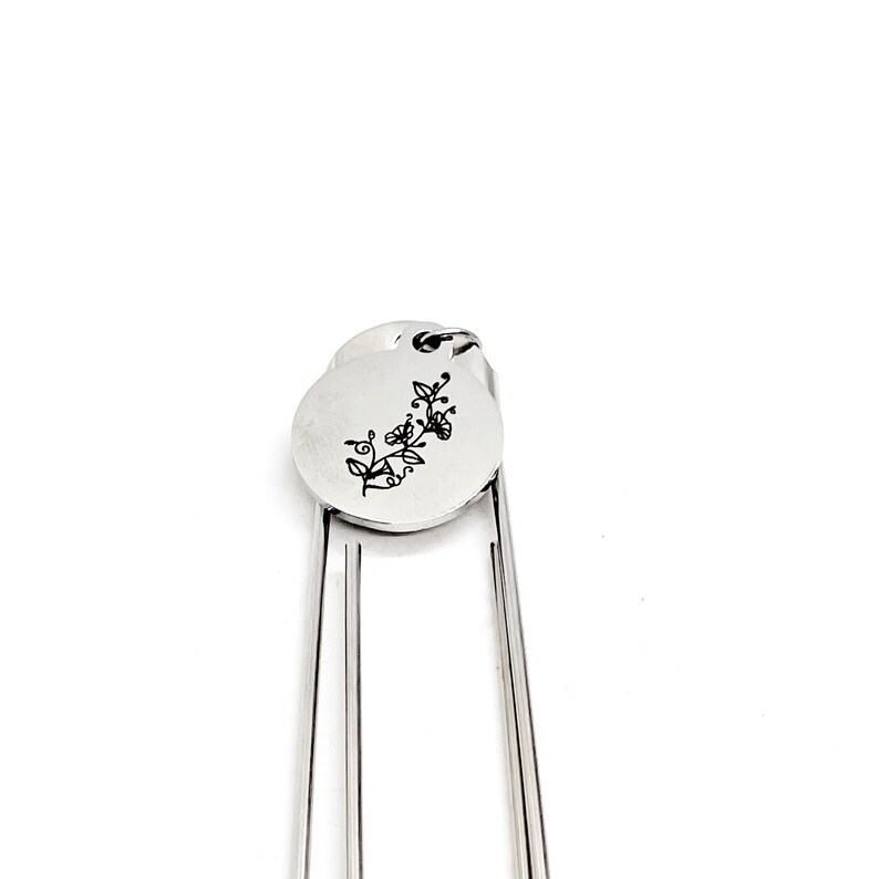 May include: Silver bookmark with a round charm featuring a floral design and the word "love" engraved on it.