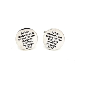 May include: Two silver-colored, round cufflinks. Each cufflink has the text "As iron sharpens iron so one person sharpens another. Proverbs 27:17" in black lettering. The cufflinks are on a white background.