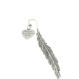 May include: A silver-colored metal bookmark with a feather design. A heart-shaped charm is attached, engraved with the words "Missing you as I walk down the aisle". The bookmark is designed to be inserted into a book.