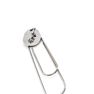 May include: Silver bookmark with a round, engraved charm featuring a floral design. The charm is attached to a silver paperclip.