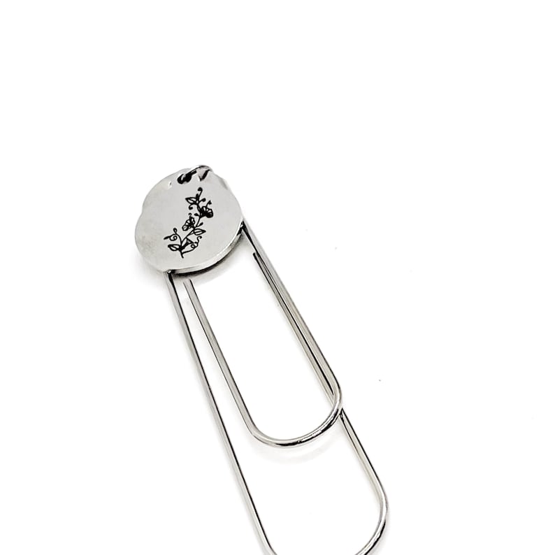 May include: Silver bookmark with a round, engraved charm featuring a floral design. The charm is attached to a silver paperclip.