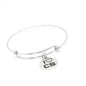 May include: A silver bangle bracelet with a circular charm. The charm is inscribed with the words "I ♡ CS". The bracelet is made of a thin, flexible metal and is designed to expand and contract.