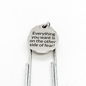 May include: A silver bookmark featuring a circular charm with the text "Everything you want is on the other side of fear!" in black. The bookmark is set against a white backdrop.