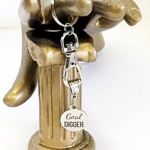 May include: Silver keychain with a silver clasp and a round silver tag that says "Goal Digger". The keychain is hanging from a gold decorative hand.