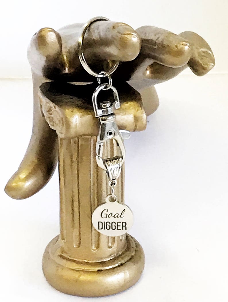 May include: Silver keychain with a silver clasp and a round silver tag that says "Goal Digger". The keychain is hanging from a gold decorative hand.