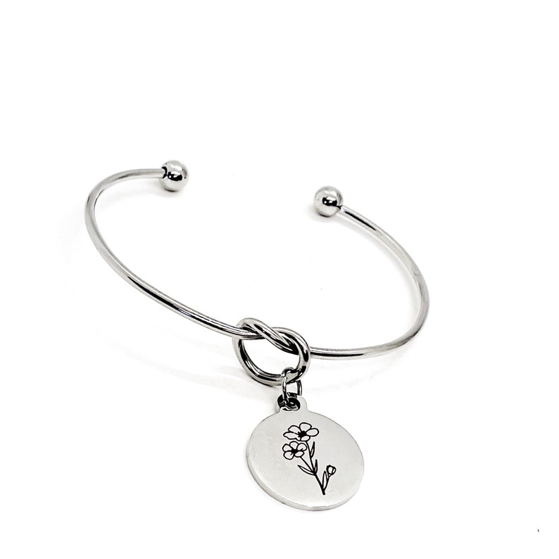 May include: A silver bangle bracelet with a knotted design and a charm featuring a delicate flower etched on a round disc.
