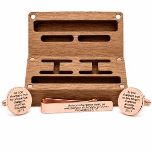 May include: A set of rose gold-toned metal cufflinks and a matching tie clip, each engraved with the text 'As iron sharpens iron, so one person sharpens another. Proverbs 27:17', presented with an open rectangular wooden storage box.