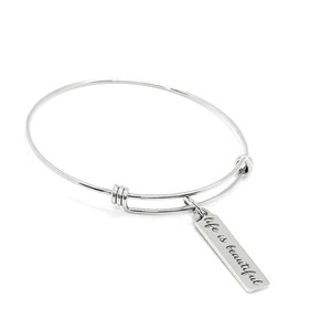 Puede incluir: Una pulsera de brazalete plateada con un dije rectangular. El dije está grabado con las palabras "life is beautiful" en una fuente cursiva. La pulsera es ajustable y tiene un diseño simple y elegante.