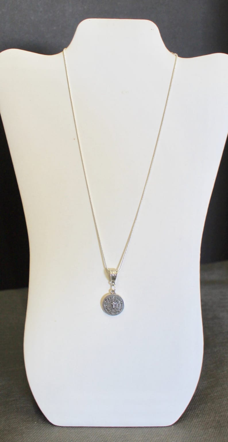 May include: A silver chain necklace with a round pendant featuring a cross and the words "Pray for us" inscribed around it.