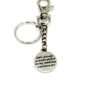May include: Silver-colored keychain with a round charm. The charm has the text "God's strength is made perfect in my weakness. 2 Corinthians 12:9". The keychain includes a clasp, a chain, and a key ring.