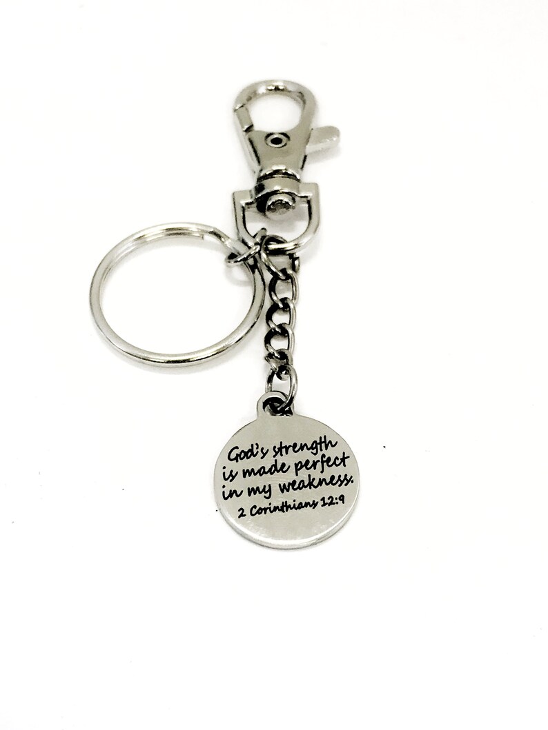 May include: Silver-colored keychain with a round charm. The charm has the text "God's strength is made perfect in my weakness. 2 Corinthians 12:9". The keychain includes a clasp, a chain, and a key ring.