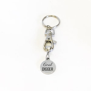 May include: Silver keychain with a silver clip and a round silver charm that says "Goal Digger" in black lettering.