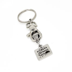 May include: Silver keychain with a silver ring and a silver clasp. The keychain has a square tag with the text "I'm into fitness taco in my mouth".
