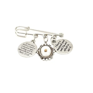 May include: A silver safety pin with three charms. One charm has a mustard seed inside a silver frame. The other two charms have inspirational quotes: "If you have faith as small as a mustard seed... nothing will be impossible for you. Matt. 17:20" and "I have a mustard seed and I'm not afraid to use it."