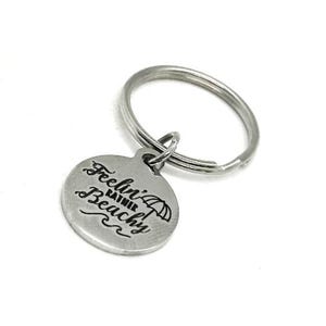 May include: A silver-colored keychain with a round charm. The charm has the phrase "Feelin' Rather Beachy" engraved in a script font, along with an umbrella and wave graphic. The keychain ring is also silver.