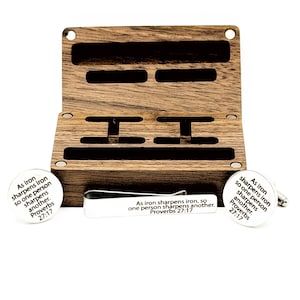 May include: A wooden box set with a tie clip and cufflinks. The box is open, revealing compartments for the accessories. The cufflinks and tie clip are white with the text "As iron sharpens iron, so one person sharpens another. Proverbs 27:17."
