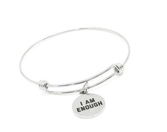 May include: A silver bangle bracelet with a circular charm. The charm features the words "I AM ENOUGH" in black text. The bracelet is set against a white backdrop.