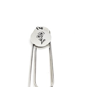 May include: Silver bookmark with a flower design engraved on a round charm. The bookmark is made of metal and has a simple, elegant design.