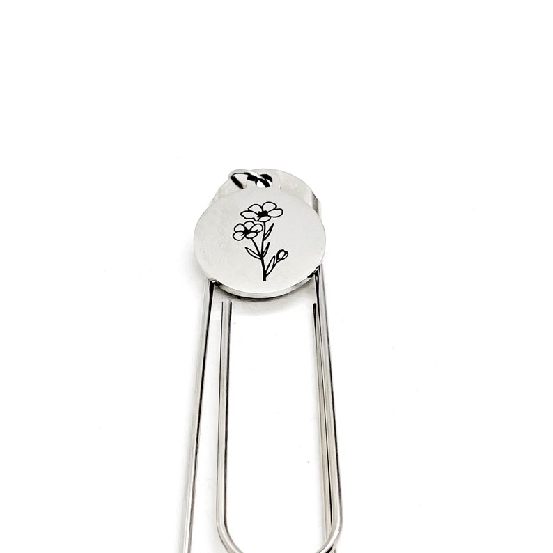 May include: Silver bookmark with a flower design engraved on a round charm. The bookmark is made of metal and has a simple, elegant design.